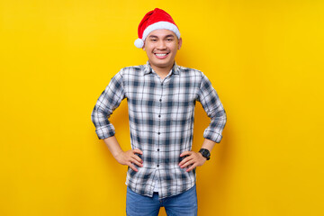 Smiling young Asian man wearing a plaid shirt in a Christmas hat posing holding hands on his waist and confident at the camera on yellow background. celebration Christmas holiday and New Year concept