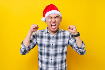 Smiling young Asian man wearing a plaid shirt in a Christmas hat posing doing winner gesture clenching fists on yellow background. celebration Christmas holiday and New Year concept