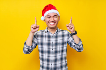 Fototapeta na wymiar smiling young Asian man wearing a plaid shirt in a Christmas hat pointing finger overhead on the workspace area on yellow background. celebration Christmas holiday and New Year concept