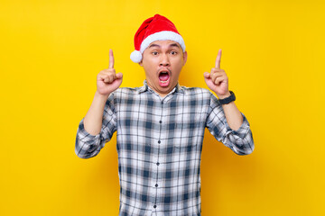 Fototapeta na wymiar Surprised young Asian man wearing a plaid shirt in a Christmas hat pointing finger overhead on the workspace area on yellow background. celebration Christmas holiday and New Year concept