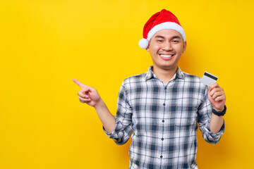 Fototapeta na wymiar Smiling young Asian man wearing Christmas hat holding credit bank card pointing index finger aside on copy space area on yellow background. celebration Christmas holiday and New Year concept