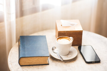 Close-up shot. Latte coffee in white cup with old book and smartphone on wooden table near bright window.