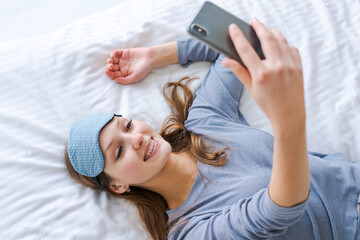 Beautiful woman in blue pajamas looking into phone. Morning starts with phone. Girl in sleep mask in bright spacious room. Reading news from phone. Attractive girl holding a phone and looking at it