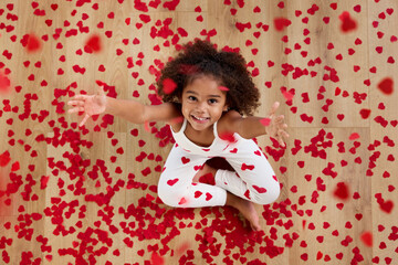 Top view of smiling young girl sitting on floor throwing heart shaped confetti