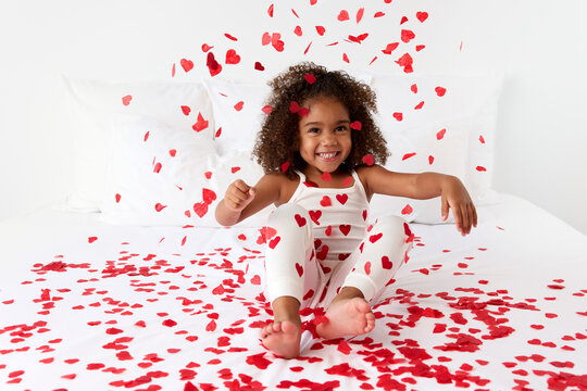 Smiling young girl sitting on bed throwing heart shaped confetti in the air