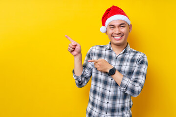 Fototapeta na wymiar Cheerful young Asian man in Santa hat pointing fingers aside at blank space for advertising text on yellow studio background. celebration Christmas holiday and New Year concept