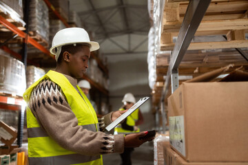 An African-American woman is working in a distribution warehouse taking inventory and moving packages. She is scanning the boxes with a barcode reader.