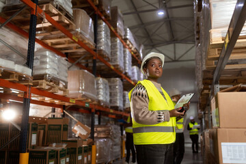An African-American woman in protective workwear is working in a distribution warehouse taking inventory and moving packages.