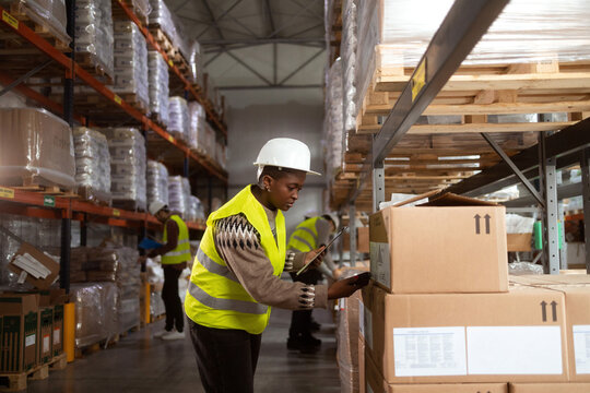 An African-American woman in protective workwear is working in a distribution warehouse taking inventory and moving packages.. She is scanning the boxes with a barcode reader.