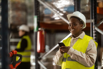 An African-American warehouse worker is using a mobile phone at work with a big smile on her face.