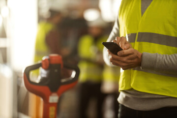 An unrecognizable warehouse worker in protective workwear is using a mobile phone at work.