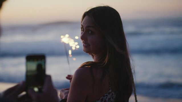 Phone, beach and sparkler with a woman recording by the sea or ocean for a new year celebration countdown. Couple, sunset and fireworks with a young female on the coast filming a video in summer
