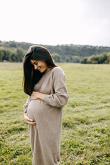 Pregnant woman with long brunette hair, holding hands on belly, outdoors, wearing a warm wool dress.