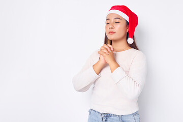 Hopeful young Asian woman in Christmas hat keeps palms pressed together in praying gesture isolated on white background