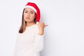 sad young Asian woman in a Santa Claus hat pointing thumb at copy space isolated over white background. celebration Christmas holiday and New Year concept