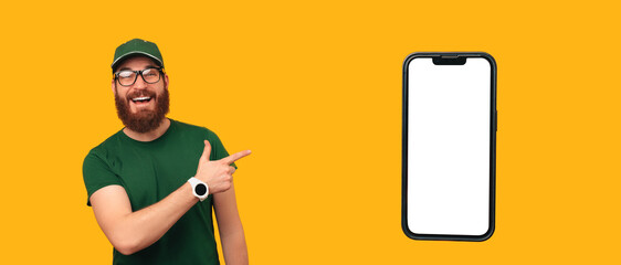 Bearded delivery man wearing green uniform points at a big phone mock up with copy space.