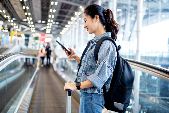 Asian Woman Tourist In Casual Clothes Using A Smartphone On Escalator At Airport Terminal. New Normal Travel At Airport