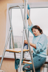 Happy Shy Mature Woman Cleaning Window. Caucasian Woman Middle Age. Elderly Woman Cleaning Apartment. Active Seniors Concept. Housewife. Concept Of Household And People In Retirement.