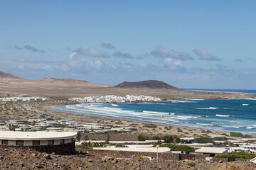 Fototapeta na wymiar View of Caleta de Famara from the bungalows. Houses. Desertic landscape. Beach with waves, village and mountains in the background. Clear sky. Famara Beach, Lanzarote, Canary Islands, Spain.
