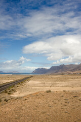 Desertic landscape. Lone black road runs through a desert plain. Mountains in the background. Blue sky with large white fluffy clouds. Yellow sand dunes with desert plants. Famara Beach, Lanzarote