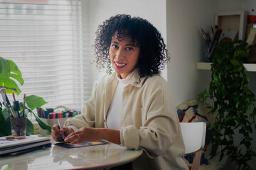 Creative artist diverse bi racial woman with curly hair in white room home studio by window sitting at desk looking and smiling at camera, surrounded by art tools, plants and shelf