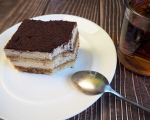 on a round white plate is a piece of tiramisu cake with a small spoon next to a glass of green tea on a dark wooden table.  side view.  dessert