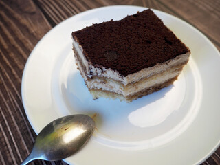 on a round white plate lies a piece of tiramisu cake with a small spoon on a dark wooden table.  side view