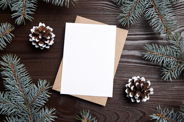 Fototapeta na wymiar Christmas card mockup with envelope, green fir tree branches and cones on brown wooden background, top view, flat lay. Blank holiday card with winter decoration