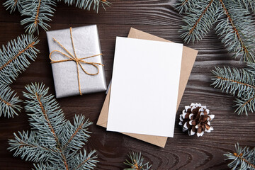 Fototapeta na wymiar Christmas greeting card mockup with envelope, gift box, green fir tree branches and holiday decorations on brown wooden background, top view, flat lay. White New Year card with decor