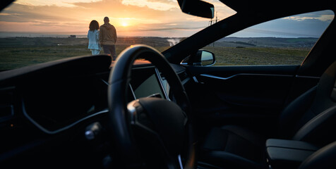 Plakat Beautiful view from automobile of young woman and man looking at orange evening sky during sunset. Snapshot of couple enjoying beauty of sundown with car steering wheel on foreground.