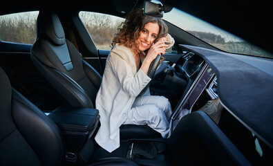 Portrait of beautiful curly lady inside electric car interior who putting arms on steering wheel. Smiling girl with curly hair adoring her automobile.