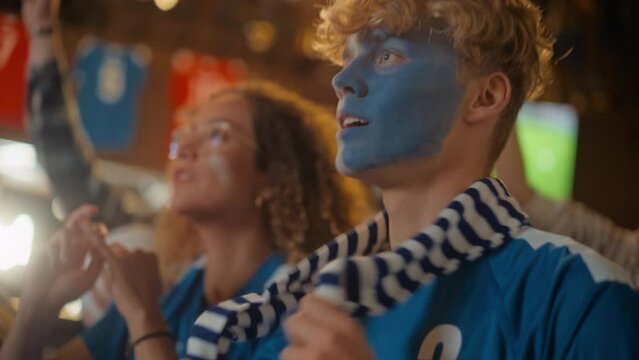 Close Up on a Group of Supportive Soccer Fans with Painted Blue and White Faces Standing in a Bar, Cheering for Their Team. Raising Hands and Shouting. Friends Celebrate Victory After the Goal.