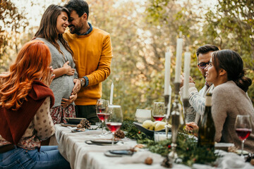 Pregnant woman and a man having an announcement to their friends during a garden party, and showing an engagement ring to them. A loving couple expecting a baby and sharing news with friends.