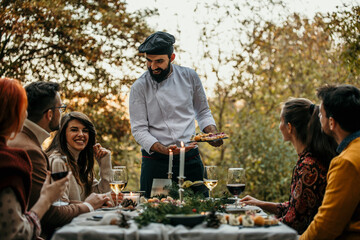 A male chef in uniform is serving a private elegant dinner to a group of people, People are smiling and welcoming the chef who is serving and bringing delicious food.
