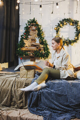 Young pretty woman with blond long hair in cozy clothes with coffee in hands using laptop watching movie and smiling on bed in room with Christmas tree at home. New Year holidays.