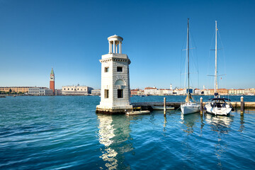 Fototapeta na wymiar Marina and lighthouse tower on San Giorgio di Maggiore with city of Venice behind. Venezia, Italy, Europe. Sailing boats, yachts moored by pier. Sunshine, daylight, calm sea water with reflections.