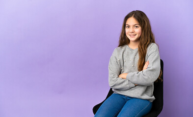 Little caucasian girl sitting on a chair isolated on purple background keeping the arms crossed in...