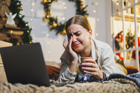 Pretty woman with blond hair in cozy clothes with coffee in hand using laptop working remotely and smiling on bed in room with Christmas tree at home. New Year holidays.