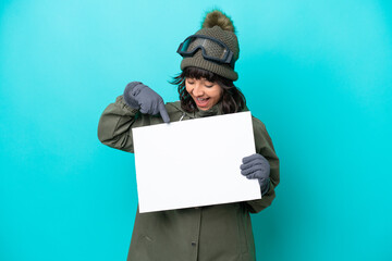 Skier latin woman with snowboarding glasses isolated on blue background holding an empty placard with happy expression and pointing it