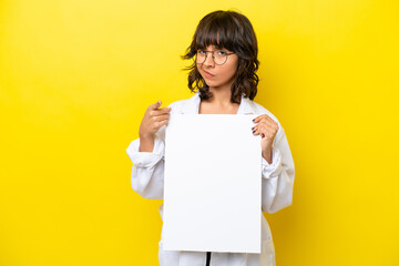 Young doctor latin woman isolated on yellow background holding an empty placard and pointing to the front