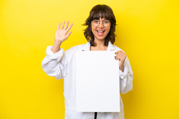 Young doctor latin woman isolated on yellow background holding an empty placard and saluting