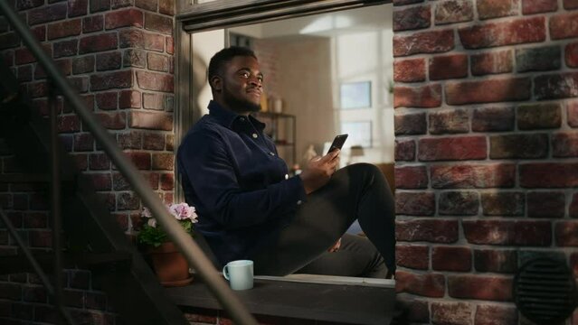 Black Male in Comfortable Casual Clothes Using Smartphone and Sitting on his Windowsill. Handsome Young Man Browsing Internet and Smiling While Watching Funny Videos and Commenting on a Website