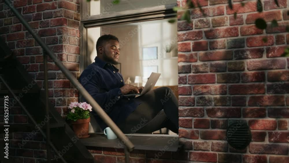 Aufkleber black young male smiling and using laptop while sitting on a windowsill. african american businessma