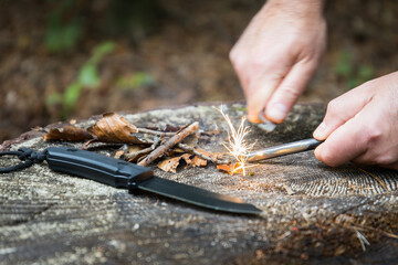 Men with knife hand starts fire with a fire steel in the woods - obrazy, fototapety, plakaty