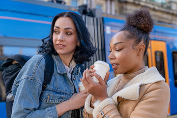 two women in a denim jacket is talking to each other, laughing, drinking coffee  and waiting for a tram at the stop Lifestyle photo