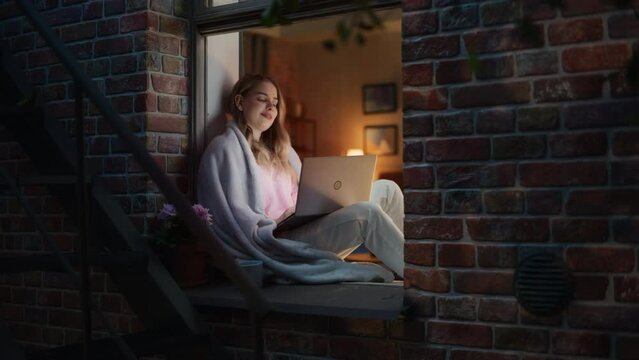 Young White Woman Using a Laptop and Covered in a Fleece blanket as She Sits on by her Window. Female Student Studying in University Abroad is Connecting to Family and Friends Through the Web