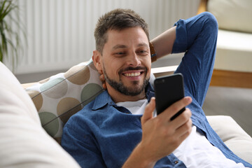 Happy man laying on sofa and using smartphone at home