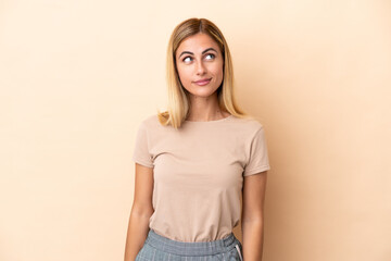 Blonde Uruguayan girl isolated on beige background having doubts while looking up