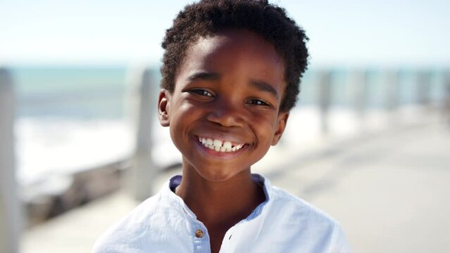 Face of happy, boy child and smile at beach promenade on summer vacation, holiday by the sea and having fun outdoors. Freedom, peace and happiness on healthy ocean trip for African children on break