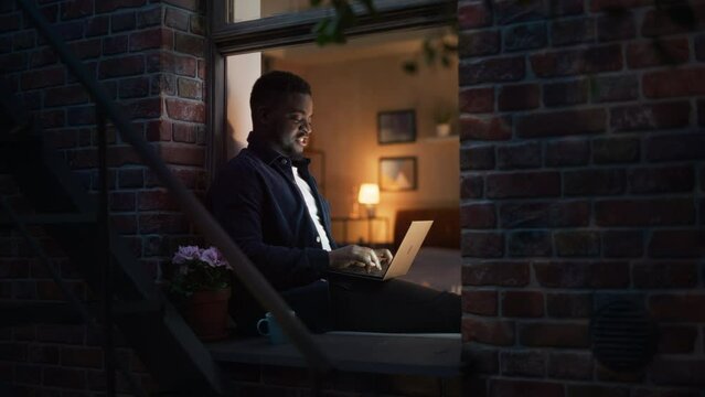 Handsome, Smiling, Black African Man Works on a Laptop While Sitting at Windowsill at Home. Portrait of a Young Male Freelancer Working on a Computer and Enjoying his Time, Making Progress on Project